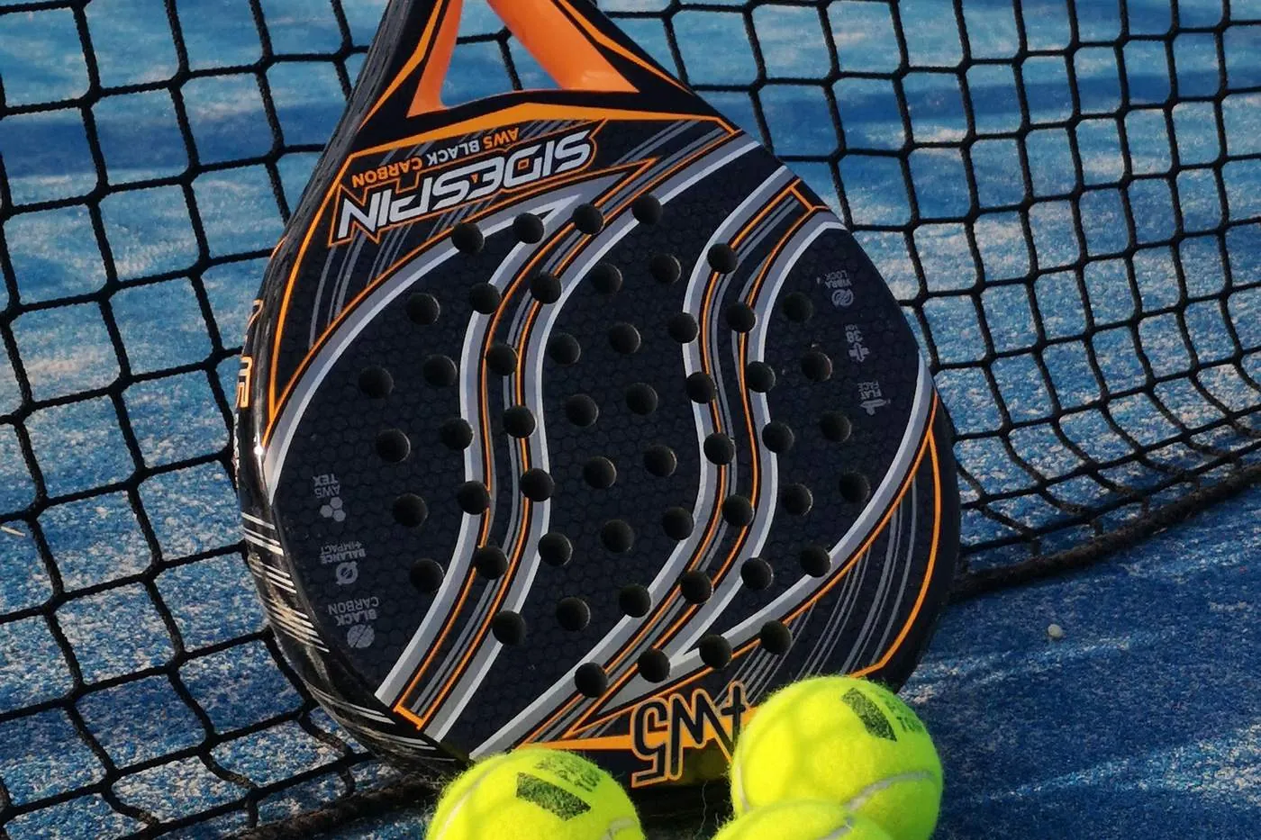 A padel racket and three balls resting against the net on a blue court